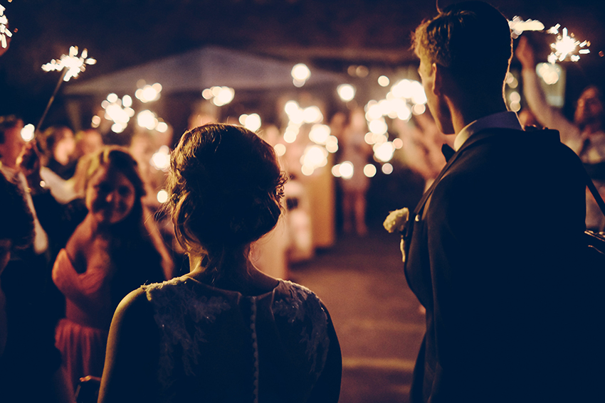 Ett bröllopspar står med ryggen mot kamera, framför sina bröllopsgäster som håller i tomtebloss som lyser i mörkret.